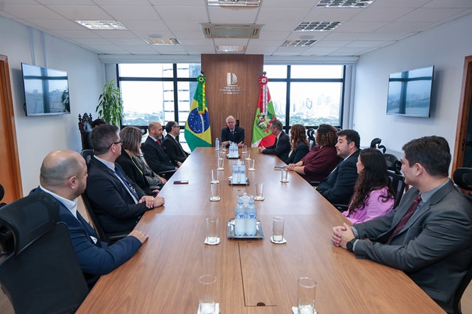 Imagem horizontal mostra desembargador Rubens Schulz reunido com autoridades, entre as bandeiras do Brasil e de SC.