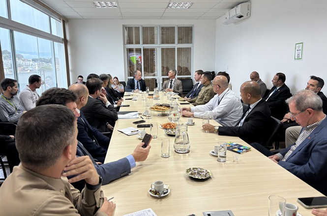 Foto horizontal: autoridades participam de reunião institucional ao redor de mesa em sala com vista para a cidade.