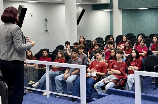 Foto horizontal: estudantes acompanham apresentação em auditório, com palestrante em pé à frente do grupo.