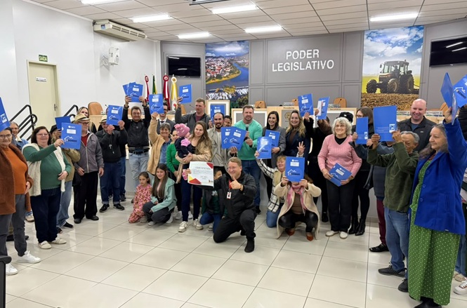 Foto horizontal: grupo posa no plenário do Poder Legislativo, exibindo livros e materiais recebidos durante atividade coletiva.