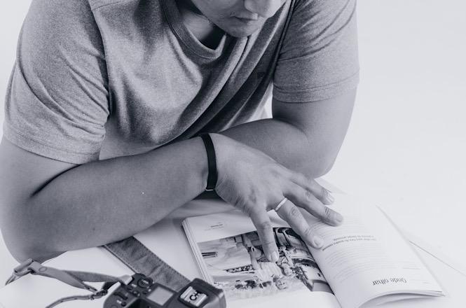 Foto horizontal em preto e branco: fotógrafo mostra um livro de fotografia aberto, com câmera fotográfica apoiada sobre a mesa.