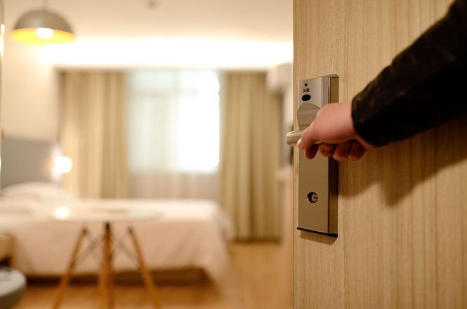 Foto horizontal: mão abre a porta de um quarto, com cama e mobiliário ao fundo.