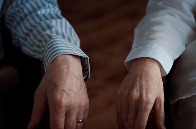 Foto horizontal: detalhe das mãos de duas pessoas posicionadas lado a lado, uma delas usando aliança.
