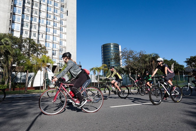 Foto horizontal: grupo de ciclistas pedala por via urbana durante passeio coletivo.