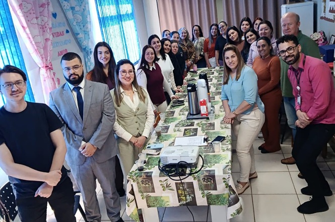 Foto horizontal: grupo de pessoas posa ao redor de mesa durante encontro em ambiente decorado, com projetor e materiais sobre a mesa.
