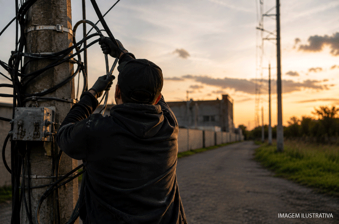 Foto horizontal ilustrativa: trabalhador manuseia cabos instalados em poste de energia elétrica, em via urbana ao entardecer.