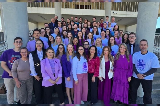 Foto horizontal: grupo de homens e mulheres posa para registro coletivo em escadaria, com roupas e camisetas em tons de roxo, alusivas a uma campanha.
