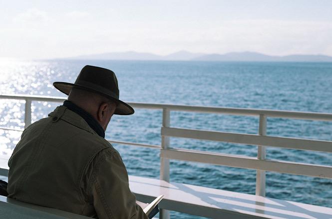 Foto horizontal: homem de chapéu lê sentado em embarcação, com o mar e montanhas ao fundo.