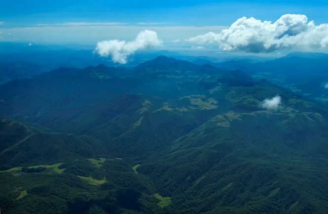 Foto horizontal: vista aérea de extensa região montanhosa coberta por vegetação, sob céu azul e nuvens.