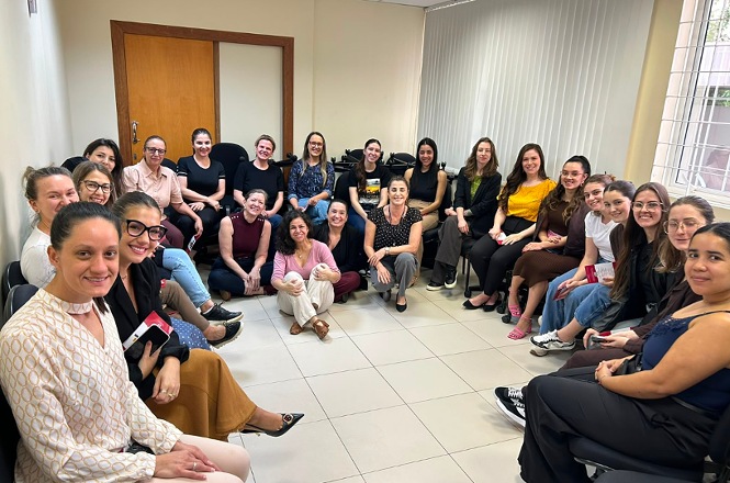 Foto horizontal: grupo de mulheres posa sorridente para registro coletivo, reunido em círculo em uma sala.