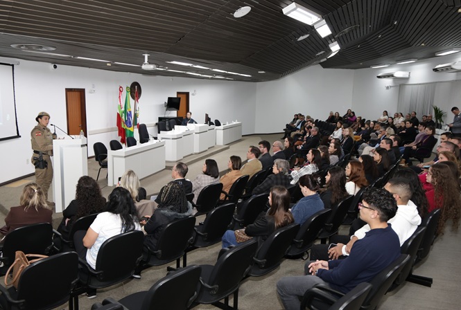 Foto horizontal: policial militar fala ao microfone diante de público reunido em auditório, durante palestra.