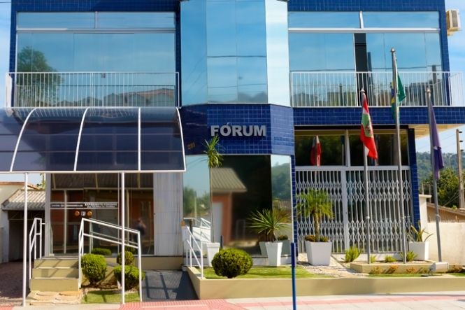 Foto horizontal: vista externa da fachada de prédio do Fórum, com bandeiras hasteadas, jardins e acesso principal em destaque.