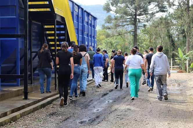 Foto horizontal: grupo de pessoas caminha por área externa arborizada, ao lado de estrutura em tons de azul e amarelo.
