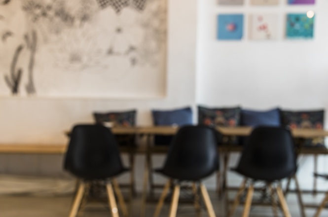 Foto horizontal: ambiente interno com mesa e cadeiras, decorado com quadros e almofadas ao fundo, em imagem com foco suave.