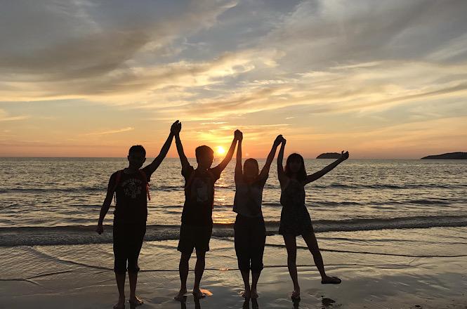 Foto horizontal: quatro pessoas posam de mãos dadas à beira-mar, com os braços erguidos, durante o pôr do sol.