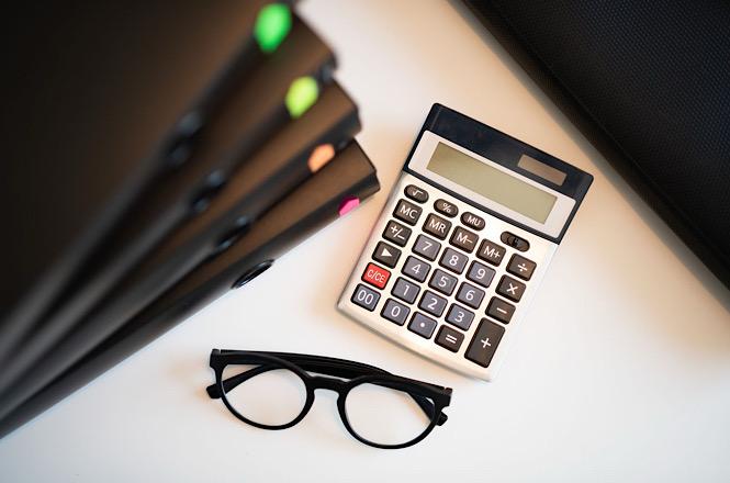 Foto horizontal: calculadora, óculos e pastas de documentos aparecem dispostos sobre uma mesa de trabalho.