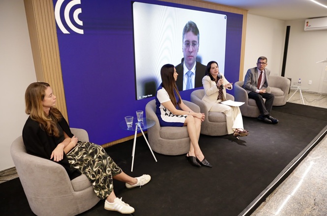 Foto horizontal: mulher fala ao microfone durante debate em painel, acompanhada por outros participantes, com homem em participação virtual exibido em telão ao fundo.