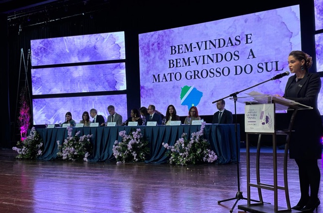 Foto horizontal: mulher discursa ao microfone durante abertura de evento, diante de mesa composta por autoridades e telão de boas-vindas a Mato Grosso do Sul.