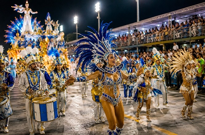 Foto horizontal: integrantes de escola de samba desfilam com fantasias em tons de azul, branco e dourado, diante do público nas arquibancadas.