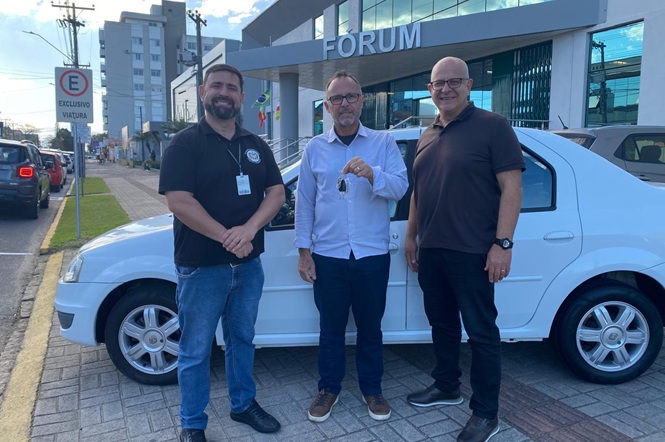 Foto horizontal: três homens posam ao lado de veículo branco em frente ao prédio do Fórum; o homem ao centro segura a chave do automóvel.