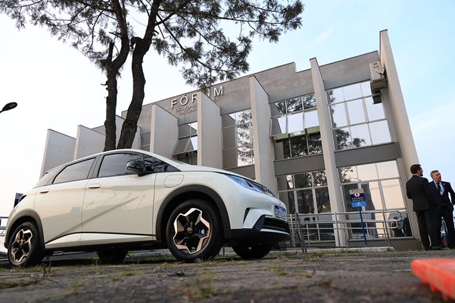 Foto horizontal: veículo elétrico aparece estacionado em frente ao prédio do Fórum, com duas pessoas ao fundo.