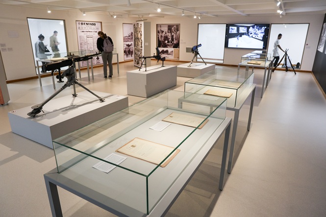 Foto horizontal: exposição histórica reúne documentos em vitrines, armamentos antigos e painéis com fotografias e registros de época.