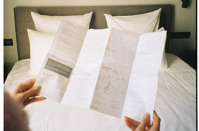 Foto horizontal: pessoa segura documentos impressos diante de uma cama, em ambiente que remete a quarto de hotel.