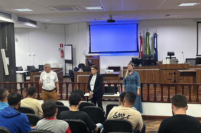 Foto horizontal: representantes de projeto social conversam com grupo de homens reunidos em auditório, diante de espaço destinado a sessões judiciais.