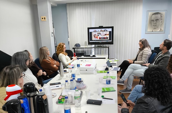 Foto horizontal: grupo de pessoas participa de reunião ao redor de mesa, com apresentação exibida em monitor ao fundo.