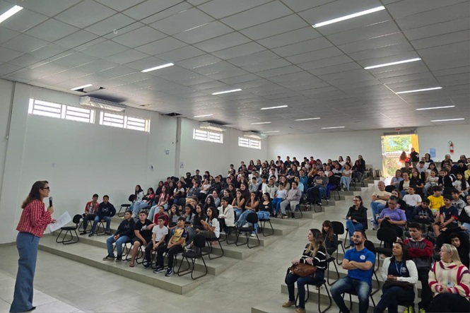 Foto horizontal: mulher fala ao microfone diante de estudantes e adultos reunidos em auditório durante atividade educativa.