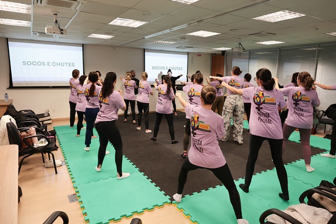 Foto horizontal: grupo de mulheres participa de atividade prática de defesa pessoal, realizando movimentos de socos e chutes em sala de treinamento.