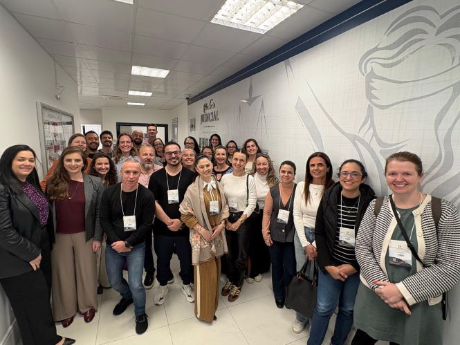 Fotografia de grupo registrada em um corredor interno de prédio institucional. Cerca de 25 pessoas estão posicionadas lado a lado e em diferentes fileiras, voltadas para a câmera. O ambiente possui piso claro, iluminação por painéis no teto e paredes brancas. Na parede à direita há um grande desenho estilizado da figura da Justiça, com venda nos olhos, e, ao fundo, uma identificação visual institucional. À esquerda aparecem quadros informativos fixados na parede. As pessoas usam crachás de identificação e vestimentas variadas, em contexto de encontro ou atividade profissional.