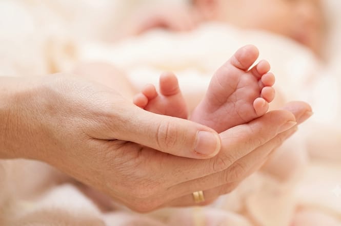 Foto horizontal: mãos de adulto seguram delicadamente os pés de um bebê, em gesto de cuidado e proteção.