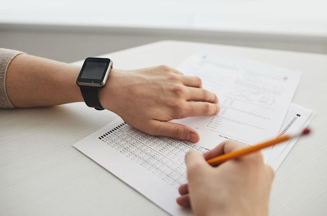 Foto horizontal: pessoa preenche formulário com lápis sobre uma mesa, enquanto mantém uma das mãos apoiada sobre os documentos.