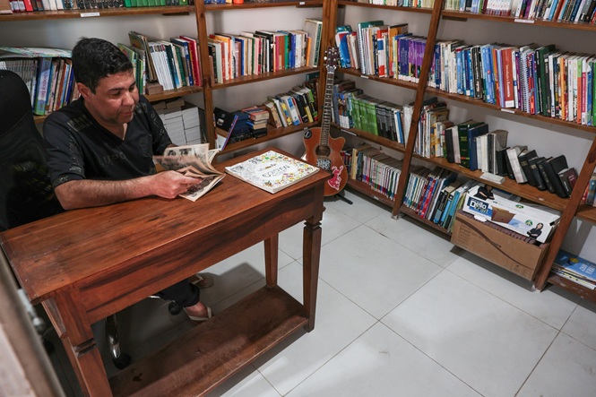 Foto horizontal: homem consulta material impresso em biblioteca, sentado à mesa cercada por estantes repletas de livros.