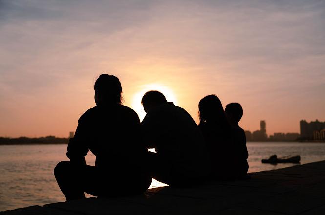 Foto horizontal: silhuetas de quatro pessoas sentadas à beira da água, reunidas durante o pôr do sol.