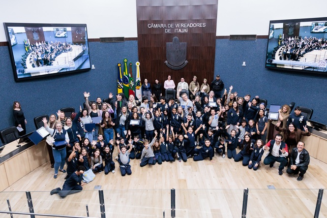 Foto horizontal: grande grupo de estudantes e profissionais posa para registro coletivo no plenário da Câmara de Vereadores de Itajaí, com braços erguidos.