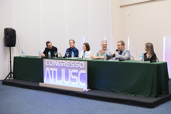 Foto horizontal: seis participantes compõem mesa de debate durante o Congresso Atjusc, com um dos integrantes falando ao microfone.
