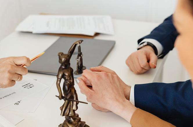 Foto horizontal: pessoas participam de reunião em torno de uma mesa, com estatueta da Justiça, documentos e materiais de trabalho.