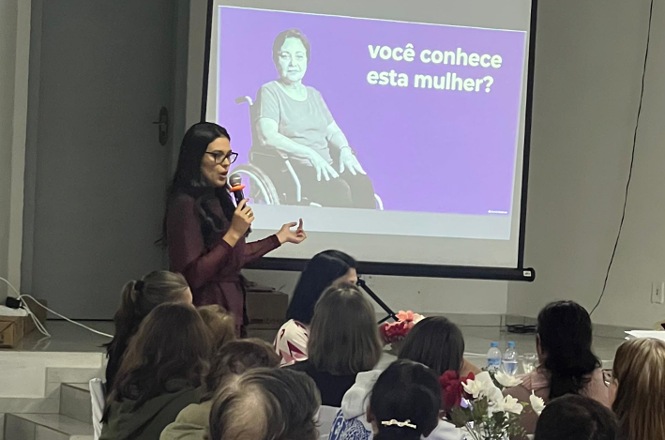 Foto horizontal: juíza Nathana Campos Dias de Souza fala ao microfone durante palestra, com imagem de Maria da Penha, em cadeira de rodas, projetada ao fundo.