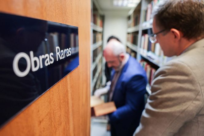Foto horizontal: placa com a inscrição “Obras Raras” em primeiro plano, enquanto dois homens consultam livros entre estantes de biblioteca ao fundo.