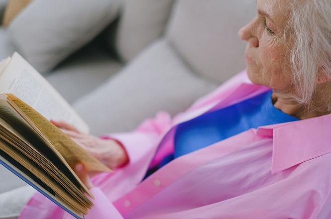 Foto horizontal: mulher idosa, de camisa rosa, lê um livro enquanto está sentada em um sofá.