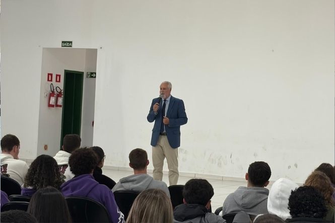 Foto horizontal: homem de terno azul fala ao microfone diante de estudantes sentados em auditório.
