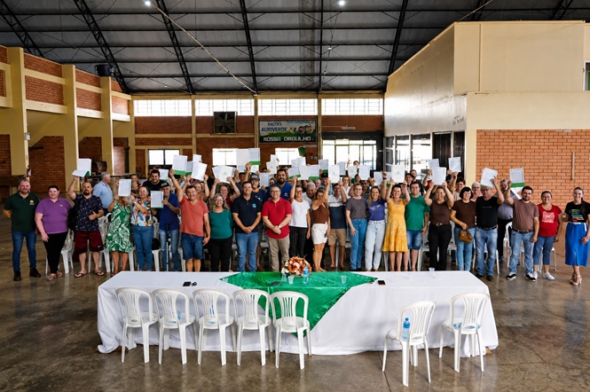 Foto horizontal: grupo de homens e mulheres posa em salão, segurando documentos, diante de mesa coberta com toalhas branca e verde.