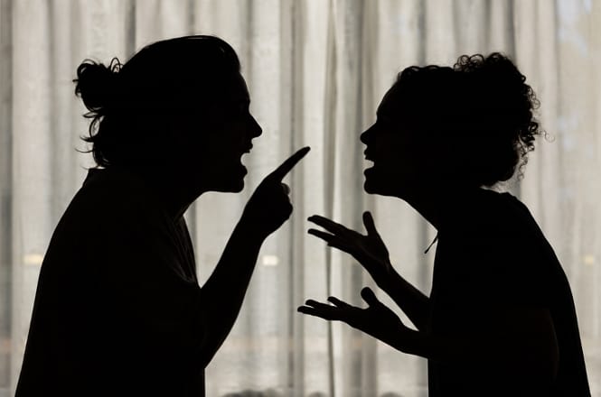 Foto horizontal: silhuetas de duas mulheres discutindo, frente a frente, com gestos de confronto.