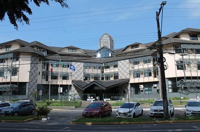 Foto horizontal: fachada de prédio do Fórum, com bandeiras em frente e veículos nas cores preta, vermelha, branca e prata estacionados.