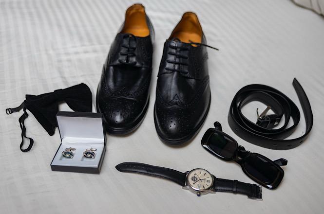 Foto horizontal: sapatos e cinto pretos, relógio, óculos escuros e abotoaduras estão dispostos sobre uma superfície branca.