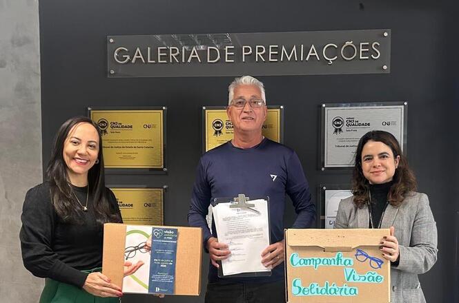 Foto horizontal: duas mulheres e um homem posam diante da Galeria de Premiações, segurando materiais da Campanha Visão Solidária.