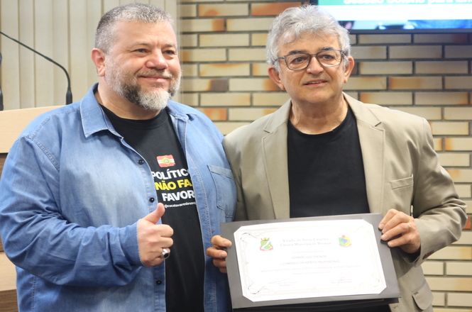 Foto horizontal: dois homens posam lado a lado; um deles segura certificado emoldurado, enquanto o outro faz sinal de positivo.