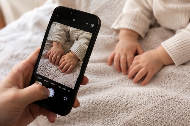 Foto horizontal: pessoa fotografa com celular as mãos de um bebê apoiadas sobre uma manta clara.